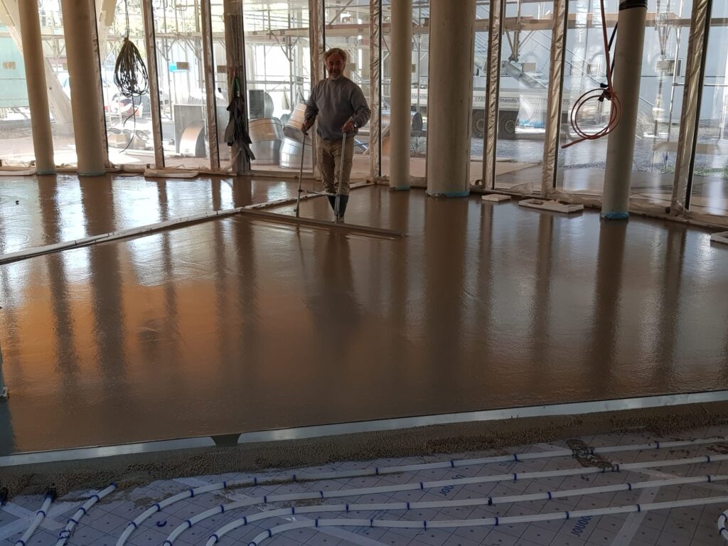 Handwerker glättet Betonboden in einem lichtdurchfluteten Raum, konzentrierter Ausdruck, Baustelle.