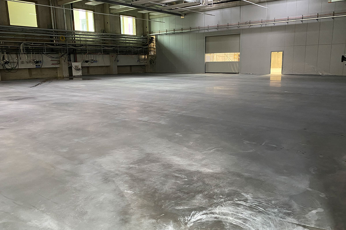 Leerer, großer, grauer Raum mit Betonboden und hohen Wänden; industrielle Atmosphäre.