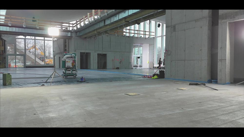 Große, leere Baustelle innen mit grauen Betonwänden und viel natürlichem Licht durch Fenster.