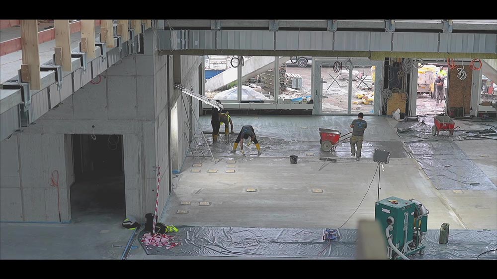 Mehrere Bauarbeiter bei der Arbeit in einer unfertigen Industriehalle mit Betonwänden.