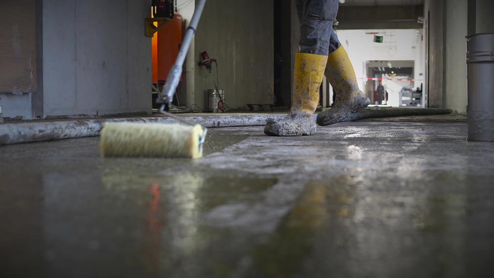 Person mit gelben Gummistiefeln streicht Betonboden grau; Arbeitsumgebung, Fokussierung auf Werkzeug.