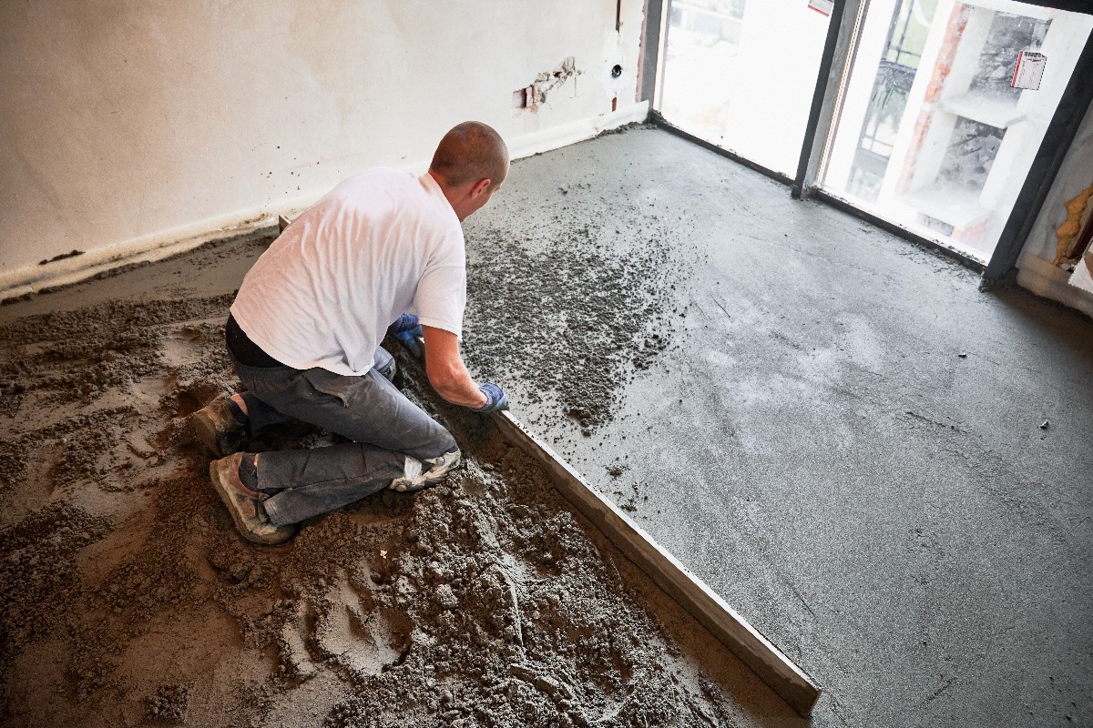 Male construction worker using screed rail while screeding floor in living room. Man flattening and smoothing surface with straight edge in apartment during renovation.